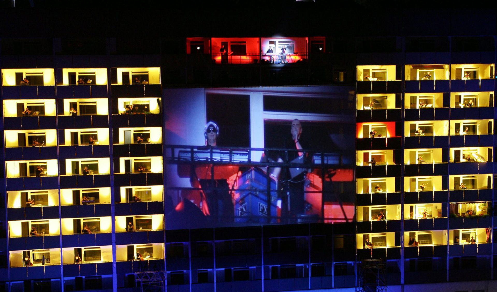 Auf den Balkonen eines Plattenbaus stehen verteilt und hell beleuchtet die Mitglieder der Dresdner Sinfoniker und spielen ihre Instrumente. In der Mitte hängt eine große Leinwand auf der die Übertragung der Sinfonie läuft.